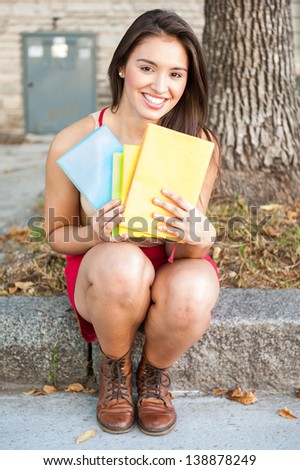 Pretty young female student with a beautiful smile sitting on a curb stone at the side of a road holding colourful folders in her hands