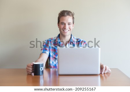 Young adult using computer and holding cup of coffee whilst looking at camera.