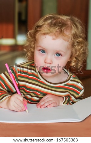 Closeup portrait of preschooler with strawberry blonde curly hairs who draws in the sketchbook by pencil and looks into the lens