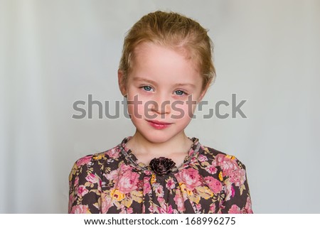 Closeup portrait of calm preschool girl with strawberry blonde hairs and flowered dress who looks into the lens against the light grey background