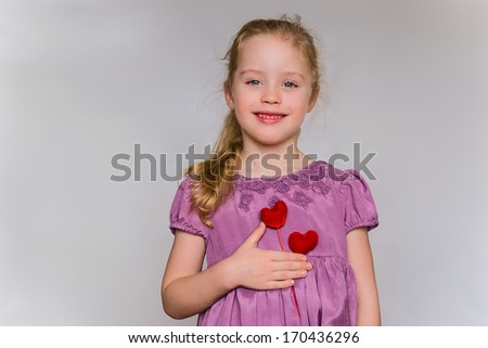 Preschool cute girl with strawberry blonde hairs and lilac dress who holds two red heart pattern on the chest against the light grey background