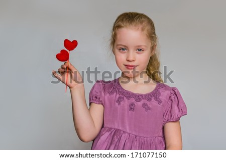 Preschool cute girl with strawberry blonde hairs and lilac dress who holds two red heart pattern against the grey background