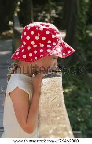 Girl in red white polka dots panama looks down from a bridge against green foliage background