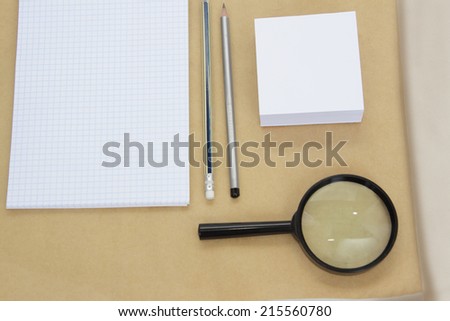 Paper notebook and magnifying glass.Notebook, pencils, paper and magnifying glass against a rough sheet of paper.
