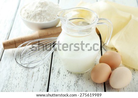 Bowl of wheat flour with eggs, whisk and glass on milk on white wooden background
