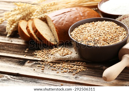 Ears of wheat and bowl of flour and wheat grains on brown wooden background