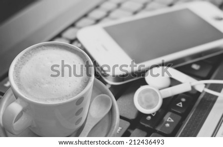 Coffee cup with smart phone and headphones on laptop, black & white style.