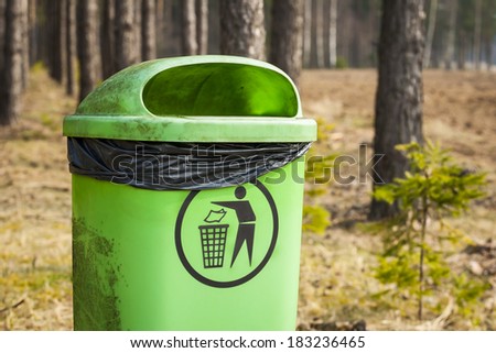 Green trash basket with sign pictogram in forest, plastic bag inside visible.