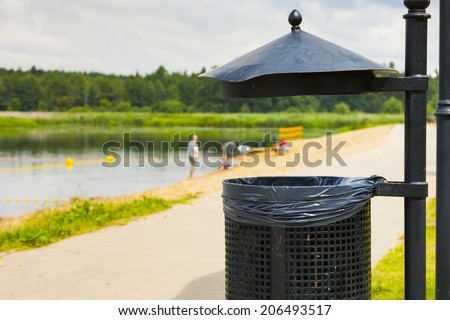 Trash basket with black plastic bag, standing in park in the background of beach. Metal roof of trash.