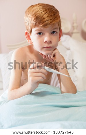 Caucasian boy with red hair sitting under the covers in his bed with a thermometer in his hand, looking sad.