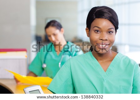 Smiling African American nurse at hospital work station lit brightly.  Second female medical professional in background.