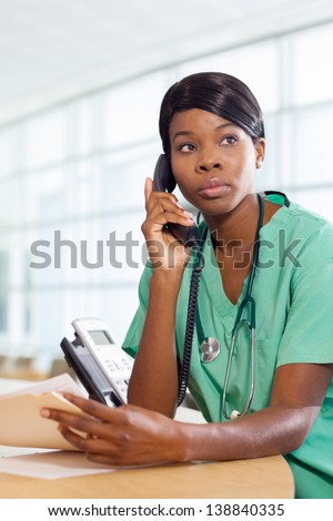 Serious African American nurse at hospital work station on the phone with a patient file and stethoscope.