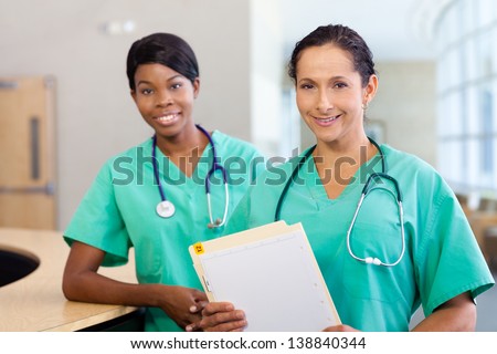 Smiling African American and caucasian nurse at hospital work station lit brightly with carts and wearing stethoscopes.  Caucasian nurse in foreground.