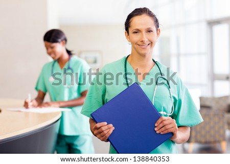 Smiling caucasian nurse at hospital work station lit brightly with phone and stethoscope.  African American nurse  in background.