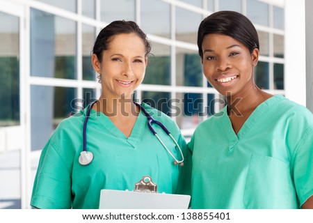 Smiling latin and afro-american nurse holding a clipboard in brightly lit exterior hospital environment in scrubs, white lab coat and holding glasses.