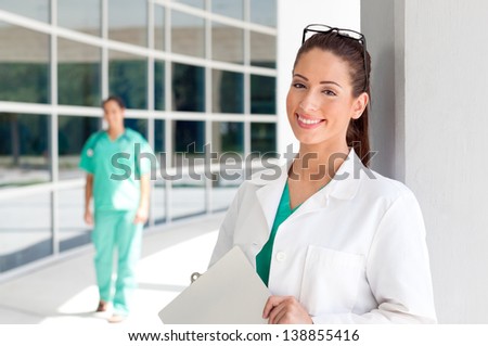Smiling Caucasian doctor holding a clipboard in brightly lit exterior hospital environment in scrubs, white lab coat. Nurse in scrubs in the background.