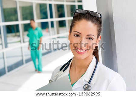 Smiling Caucasian and African American nurse and or doctor holding a clipboard in brightly lit exterior hospital environment in scrubs, white lab coat and holding glasses.