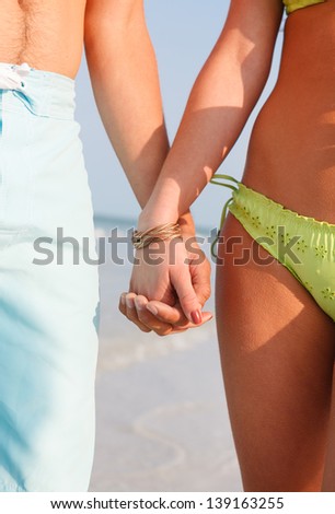Closeup of A Young Couple Holding Hands on the Beach. Woman wears green bikini. Man wears light blue shorts. Front View.