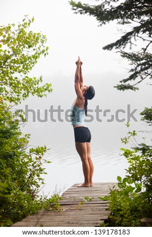 middle aged woman practicing yoga and starting half moon with hands to feet pose on dock by a lake in the foggy morning, vertical composition, Surry Maine