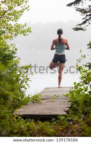 middle aged woman practicing yoga, tree pose, on dock by a lake in the foggy morning, vertical composition, Surry Maine
