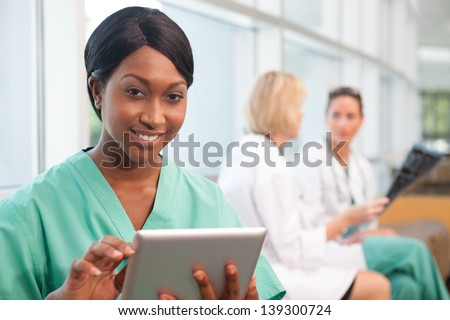 Smiling African- American nurse holding tablet with . a Caucasian and Hispanic doctor in background, looking at scan in hospital