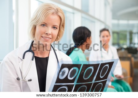 Female Caucasian doctor holding x-ray, scan, sitting with African-American nurse or doctor and Hispanic nurse talking in background, in hospital waiting area or lobby