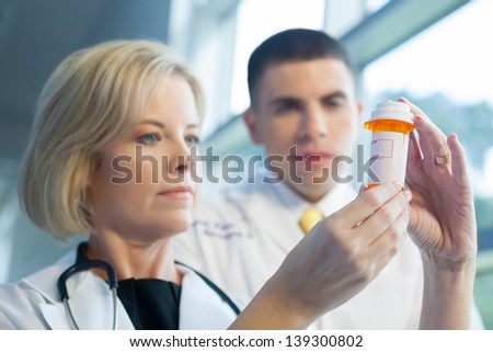 Caucasian male and female doctors in white lab coats looking at a prescription bottle in brightly lit hospital. Bottle is in selective focus.
