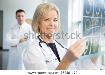 Female blonde caucasian doctor white lab coat with x-ray in brightly lit hospital smiling at camera. Caucasian male doctor in background