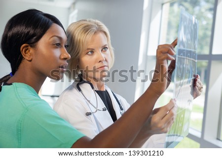 African American nurse in scrubs and Caucasian doctor in scrubs and white lab coat looking at an x-ray in brightly lit  hospital..