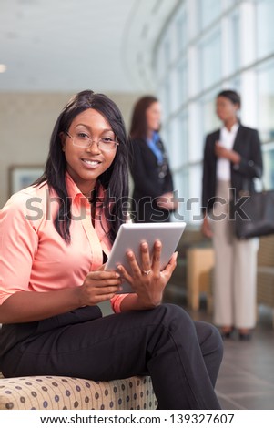 african-american business woman sitting in lobby with computer tablet, looking at camera, smiling, two business women in background