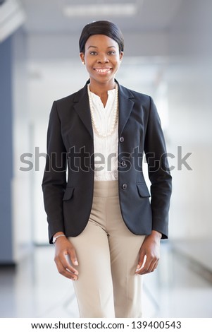 Young African-American professional woman in a hallway, cheering, arms raised, smiling, wearing blazer, pearls