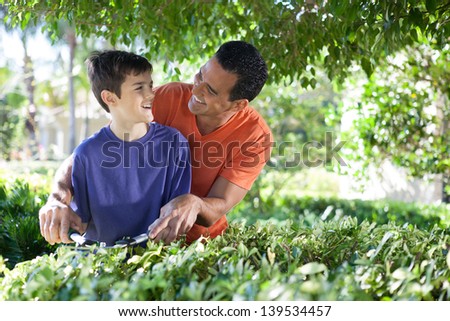 Hispanic father happily teaches his teenaged son how to use hedge clippers to trim bushes in lush yard.