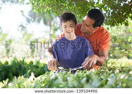 Hispanic father happily teaches his teenage son how to use hedge clippers to trim bushes in lush yard.