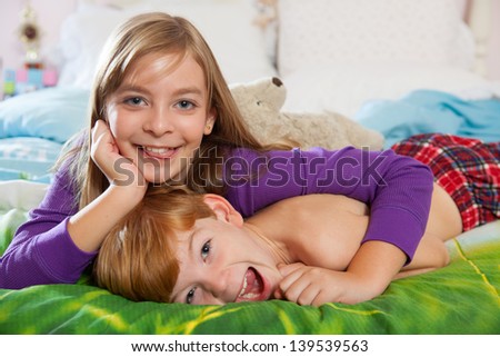 Blonde haired sister and red haired brother laughing and lying happily on bed in pajamas looking at camera.