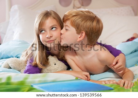 Blonde haired sister getting kissed on the cheek by red haired brother, lying on bed in pajamas.
