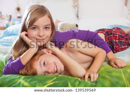 Blonde haired sister and red haired brother lying happily on bed in pajamas looking at camera.