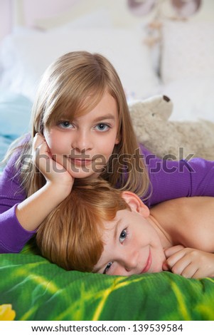 Blonde haired sister and red haired brother lying happily on bed in pajamas looking at camera.