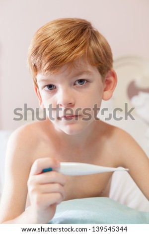 Caucasian boy with red hair sitting under the covers in his bed with a thermometer in his hand, looking sad.