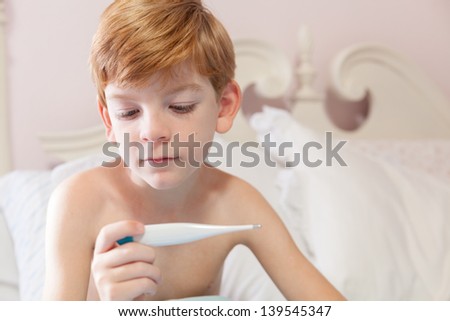 Caucasian boy with red hair sitting under the covers in his bed looking at the thermometer in his hand.