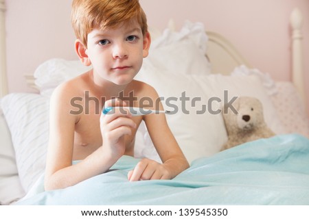 Caucasian boy with red hair sitting under the covers in his bed with a thermometer in his hand, looking at the camera.