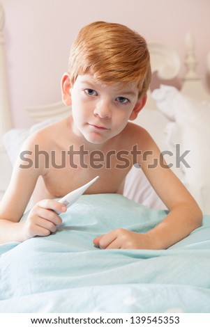 Caucasian boy with red hair sitting under the covers in his bed with a thermometer in his hand, looking at the camera,sad.
