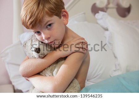 Young caucasian boy with red hair, sitting on edge of bed with sad expression, holding teddy bear, looking at camera.