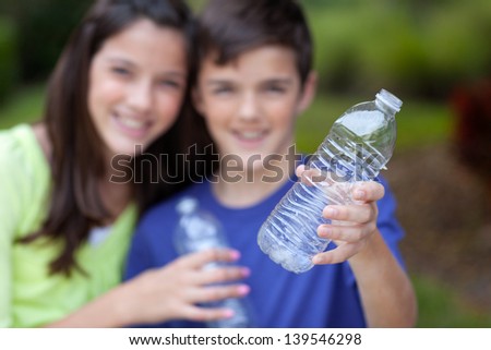 Smiling caucasian boy and girl holding up clear water bottles outside in yard, for recycling, with bottle in focus in foreground