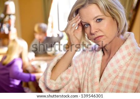 Caucasian blonde mom  in foreground looking at camera, in bathrobe holding head in frustration, while kids fight in background in kitchen.