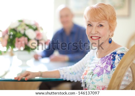 Happy caucasian Senior Couple in Dining room. Female sitting looking at camera. Senior husband out of focus looking at camera. Bright and Airy.