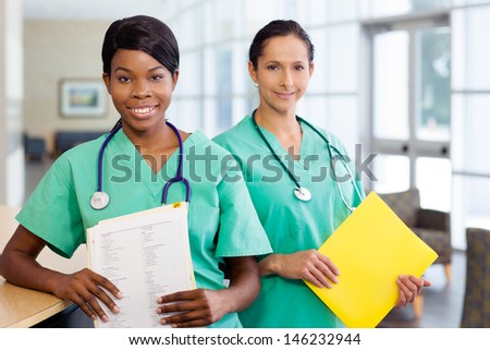 Smiling African American and caucasian nurse at hospital work station lit brightly with carts and wearing stethoscopes.