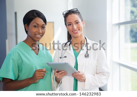 Smiling african american nurse in scrubs and caucasian doctor in scrubs and white lab coat holding a tablet computer in brightly lit  hospital halway.