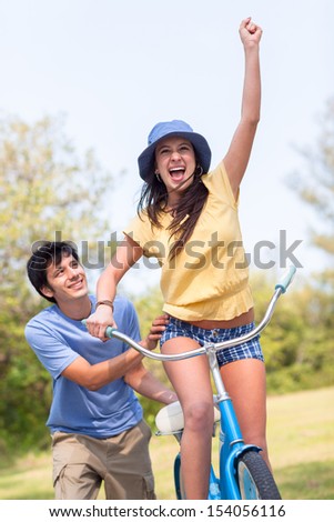 Young latin man in shorts and blue shirt helps young woman riding blue bike with green trees and grass. Horizontal shallow focused composition.