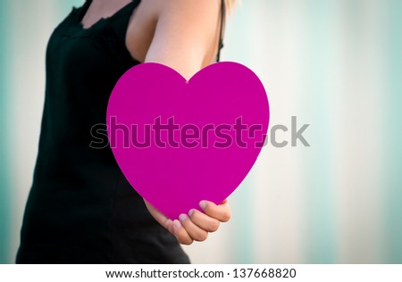 Young female walking out of the frame and boldly holding out a large magenta heart.