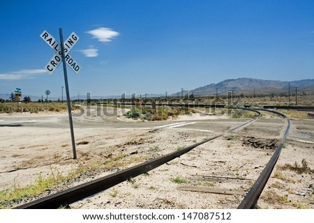 A disused railway line runs through a white desert. A railway crossing sign marks the intersection with a dirt track. The sky is deep blue with few tiny clouds.